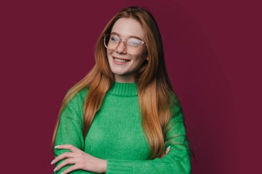 Gözlüklü, yeşil süveterli düşünceli bir kadın kestane rengi arka plan kollarına karşı gülümsüyor.