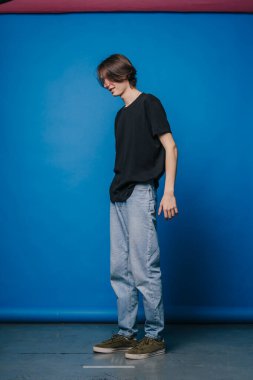Ayakta duran ve aşağı bakan genç, yumuşak stüdyo ışığı ve mavi arkaplanı olan düşünceli duruş, siyah