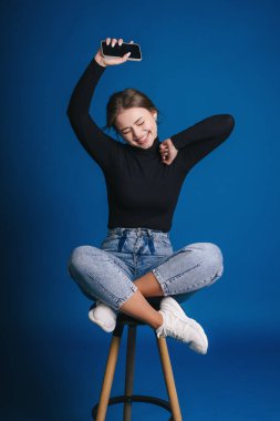 Taburede dans eden genç bir kadın, akıllı telefonu havada tutuyor, çaprazlama uyuyor, gülüyor, siyah balıkçı yaka giyiyor, kot pantolon giyiyor, beyaz spor ayakkabı giyiyor.,