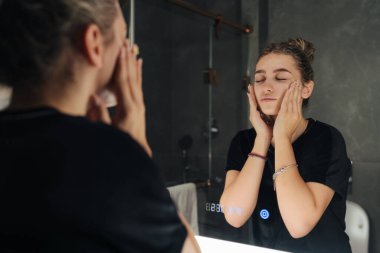 Kadın nazikçe aynada tenini kontrol ediyor, avuçlar birbirine yapışıyor ten ve dokuları değerlendirirken, yumuşak ışık ve sakin bir akşam.