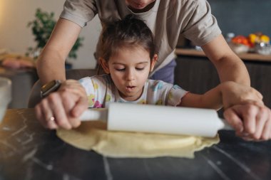 Ebeveyn ve çocuk mermer tezgahta birlikte hamuru yuvarlıyor, küçük eller yetişkin desteğiyle rulo yapıyor, un tozu.