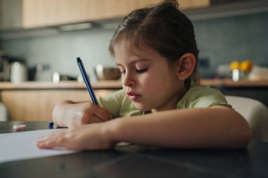 Masadaki ödevlere odaklanan genç kız, yazı yazarken kararlı duruş ve konsantre ifade, doğal mutfak arkaplanı