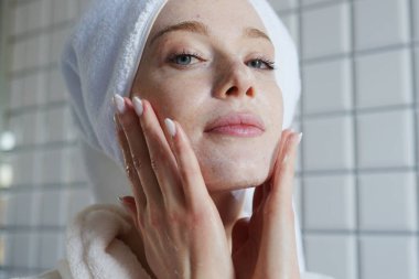 Bornozlu kadın tarafından uygulanan hidratlaştırıcı maske. Havluyla sarılmış saç, yanakları yumuşatan el kremi.