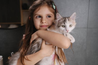 Yakın plan kız, yavru kediyi göğsüne yaslıyor, yumuşak bakışlar ve hafif ışıklandırma, saç yayı ve nötr arka plan, hassas bağ ve sakinlik.