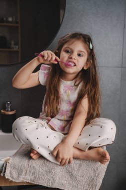 Banyoda dişlerini fırçalayan, havlunun yanında oturan, polka pijama giyen, elinde pembe bir sırıtış olan küçük bir kız.