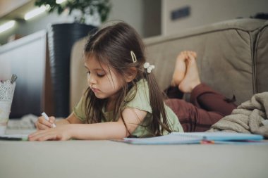 Renkli kalemlerle yere resim çizen küçük kız resimli sayfaları boyamaya ve detaylandırmaya odaklanmış, yumuşak duruş üzerine gevşemiş.