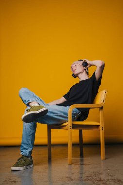 Genç adam, kulaklık takarak rahatlamış, ahşap sandalyeye oturmuş canlı sarı arka planda, gözleri kapalı, elleri başının arkasında, kot pantolon giymiş.