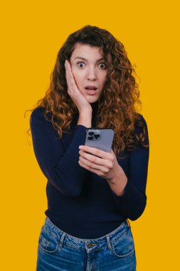 Akıllı telefonu tutan kadın şok olmuş görünüyor, elleri yanaklarında, gözleri açık, parlak sarı arka plan, samimi stüdyo