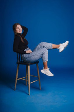 Koyu mavi arka planda kahkahalarla gülerek tabureye yaslanan genç bir kadın, rahat kot pantolon ve beyaz spor ayakkabılar.