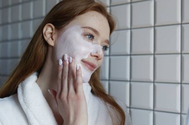 Beyaz köpük temizleyicisi yanaklara uygulanır, kadın ellerini kullanır, döşemeli banyo arkaplanı, yumuşak sabah ışığı, nazikçe...