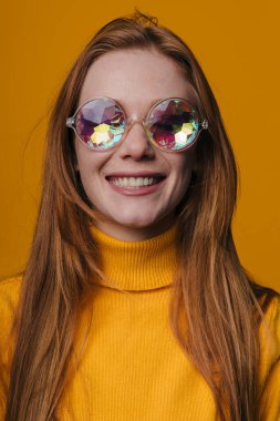 Güneş gözlüklü kızıl saçlı kadın, hardal rengi süveter, turuncu arka plan, aynalı lensler.