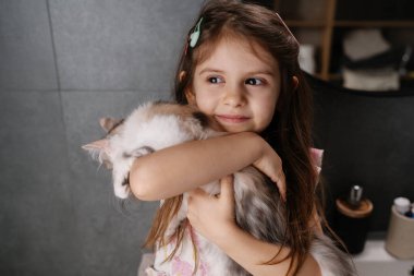 Kafkasyalı genç kız pofuduk kediye sarılıyor rahat banyo düzeninde, gülümseme, hafif aydınlatma, saç tokası,