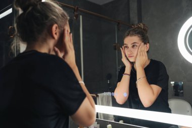 Kadın ışığın altında aynaya bakıyor, içerik yaratıcısı makyajı hazırlıyor, elleri yanaklarına bastırılmış, camda halka ışığı yansıması.,