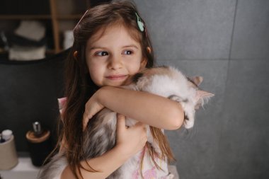 Genç kız, neşeli bir gülümsemeyle beyaz kediye sarılıyor, okuldan sonra evlat edinme günü kutlaması, sıcak, doğal bir ışıkla yakalandı, toka.