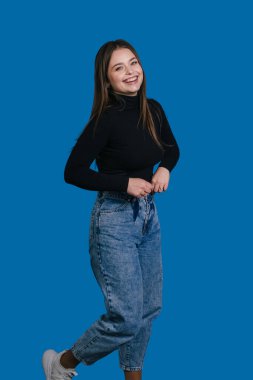 Siyah balıkçı yakalı, gülümseyen genç bir kadın canlı mavi arka planda poz veriyor, kot pantolon ve spor ayakkabılar, rahatlamış özgüven ifadesi.,