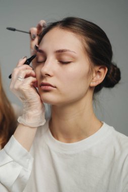 Beyaz kadın son kaş detayına kadar inceleniyor. Sanatçı, son vuruşlar için küçük fırça kullanıyor. Durgun ifade, yumuşak stüdyo.