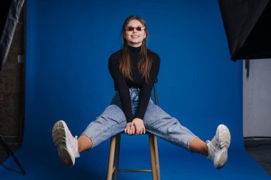 Kendinden emin beyaz kadın taburede oturuyor, güneş gözlüğü takıyor ve boğazlı kazak mavi arka planda, bacakları oyuncu bir şekilde açık.