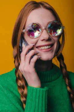 Telefondaki gülümseyen beyaz kadın, parlak stüdyo resmi, pullu kaleydoskop güneş gözlüğü, yeşil kazak, örgülü saç, sarı.