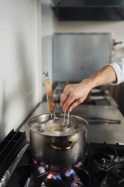 Beyaz sosis kaynamış makarna tenceresini karıştırıyor, buhar yükseliyor ve zamanlamayı ve sıcaklığı gösteren özenli duruş sergiliyor.