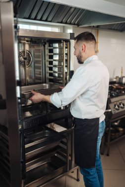 Şef ticari fırının içindeki tepsiyi kontrol ediyor, operatör rafları ve pişirme tepsilerini denetlemek için ağır fırın kapısını açıyor,