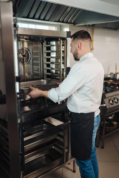 Şef ticari fırının içindeki tepsiyi kontrol ediyor, operatör rafları ve pişirme tepsilerini denetlemek için ağır fırın kapısını açıyor,