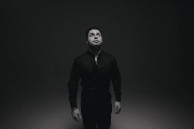 Sahne ışıkları altında siyah takım elbiseli, dik bakışlı, hala duruşu olan, iç gözlem yapan ve yüksek kontrast stüdyosunu çözen bir adam.