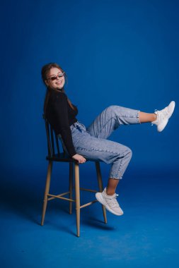 Beyaz kadın dışkı, mavi arka plan, kot pantolon ve beyaz spor ayakkabı, gözlük, şakacı bacak kaldırma, kendine güvenen gülümseme, stüdyo