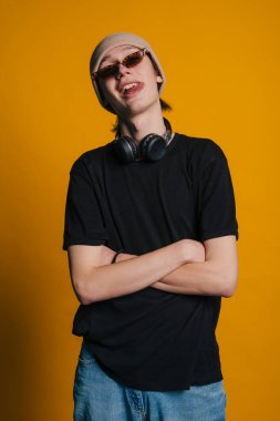 Kulaklık ve güneş gözlüklü, kolları çapraz, rahat sırıtış, siyah tişört, bere, stüdyo sarısı