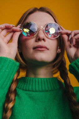 Yansıtıcı güneş gözlüğü takan, yeşil kazaklı genç bir kadın. Kaleydoskopik yansımaları, örgülü saçlarını gösteren çerçeveler kaldırmış.,