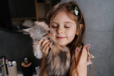 Küçük kız pofuduk kedi yavrusuna sarılıyor, gülümsüyor, gözleri kapalı, arka planda saç tokası ve kalem bardağıyla gri koltukta oturuyor, yumuşak