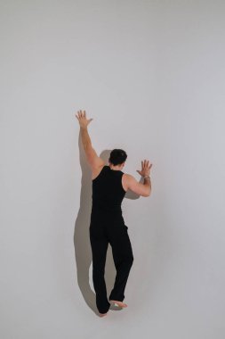 Erkek dansçı duvara yaslanıyor, dramatik gölgeler ve kontrollü poz veren minimum stüdyo ayarları, kolsuz siyah kıyafetler.,