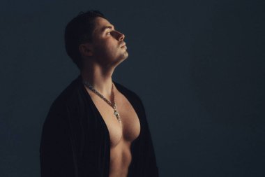 Üstsüz adam, karanlık zemine karşı poz veriyor, başı yukarı eğilmiş, kolye ışığı yakalıyor, keskin göğüs ve çene çizgisi, yumuşak.