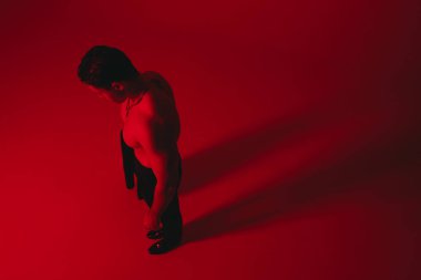 Kırmızı spot ışıkları altında üstsüz bir adam, stüdyo zemininde derin düşünceli bir poz, açık gömlek ve kravat, dramatik bir gölge.