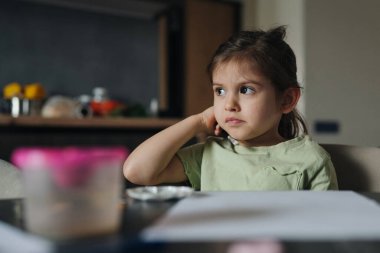 Kız, çenesini dinlendirirken düşünceli bir şekilde bakıyor. Yumuşak öğleden sonra ışığı ve iç mekan havası. Sessiz, hayal kuran ve içsel.