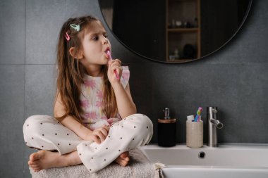 Diş fırçası, diş macunu köpüğü, pijama giysisi ve ayna yansıması ile diş fırçalayan bir kız. Kendinden emin.