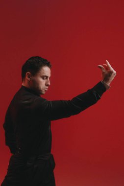 Kırmızı arka planda etkileyici erkek, narin el hareketi, odaklanmış bakış, tiyatro duruşu, sıcak yönlü ışık.,