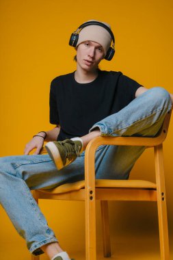 Kulaklık takan genç adam ahşap sandalyeye oturmuş, parlak sarı arka plana, bereye ve siyah tişörtlere yaslanmış.