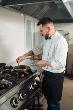 Aşçı fırının tepsisini karıştırıyor. Meşgul ticari mutfakta, kepçe gibi parlayan sos, buhar ve alev, odaklanmış duruş, gevrek.