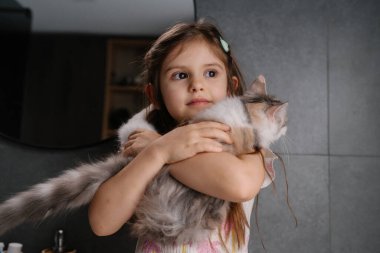 Genç kız sevgiyle uzun saçlı kediciği aynanın yanında tutuyor. Pastel elbiseli çocuk yumuşak havadaki hayvanına neşeyle sarılıyor.