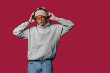 Genç beyaz adam kulaklıkları kapüşonlu ve turuncu güneş gözlükleriyle kırmızı arka plana ayarlıyor.,