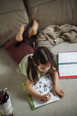 Halı çizim defterine resim çizen genç kız, işaretler dağılmış, odaklanmış ifade, sıcak oturma odası ışığı, gündelik.