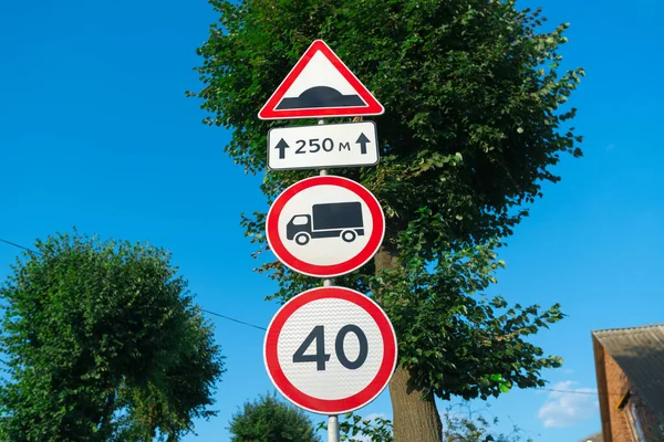 Red and white round traffic sign with black cargo truck indicating No entry for trucks speed limit 40 km/h near speed bump with trees and blue sky on the background