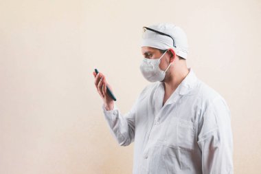 Beyaz önlüklü, koruyucu gözlüklü ve nefes maskeli genç doktor sarı arka planda fotokopi alanı olan akıllı telefonuna bakıyor..