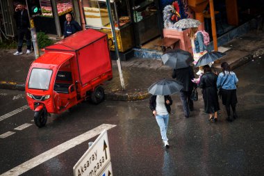İSTANBUL, Turkey. 26 Mayıs 2025. Yağmurlu bir günde, Türkiye 'nin İstanbul kentindeki ıslak bir caddeden geçen şemsiyeli yayalar, küçük bir kırmızı teslimat bisikletinin yanında.