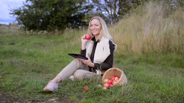 Молодая женщина ест свежее яблоко на открытом воздухе и официантский фильм на планшете — стоковое видео