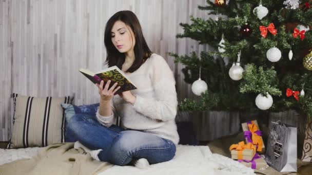 Egy gyönyörű fiatal nő ül, és Olvasson egy könyvet a karácsonyfa alatt — Stock videók