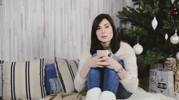 Hermosa joven sentada bajo el árbol de Navidad, bebiendo café y sonriendo — Vídeos de Stock