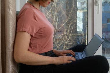 Çekici kadın elinde defter ve bir fincan çayla pencere eşiğinde oturuyor. Koronavirüs salgını sırasında evde çalışıyor.