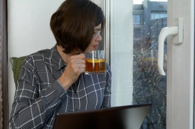 Çekici kadın elinde defter ve bir fincan çayla pencere eşiğinde oturuyor. Koronavirüs salgını sırasında evde çalışıyor.