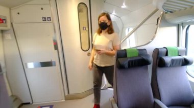 Siyah koruyucu maskeli bir kadın tren vagonuna biniyor ve koltuğuna oturuyor..